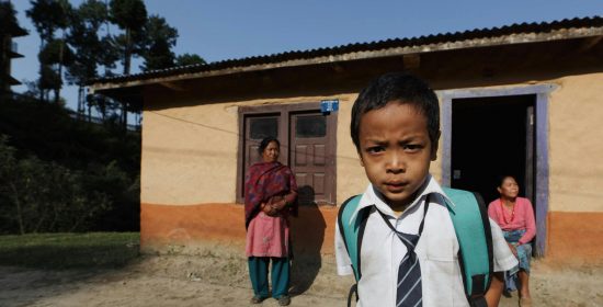 A young boy in school uniform stands in front of a rural house with two women in the background.